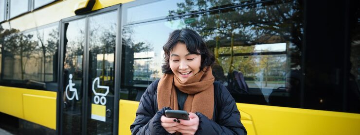 Beautiful korean girl student on bus stop looking at her smartphone checking timetable reading text