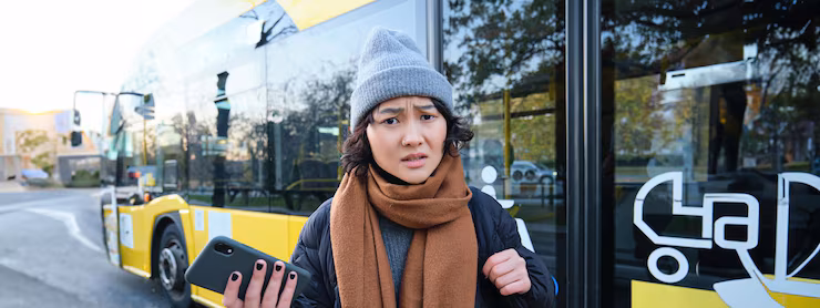 Portrait of confused asian girl standing on bus stop holding mobile phone looking shocked and upset